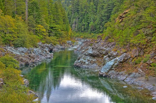 A serene river flows through a rocky landscape, surrounded by lush green trees and reflecting the sky above.