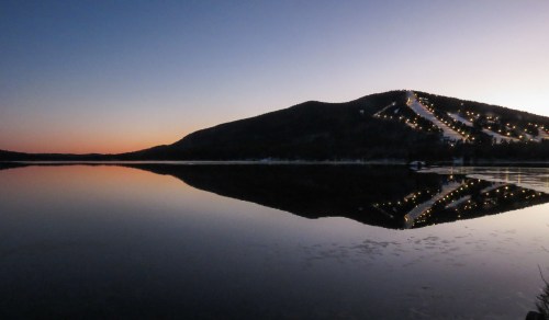 A serene lake at dusk reflects a mountain with ski slopes, illuminated by lights against a colorful sky.
