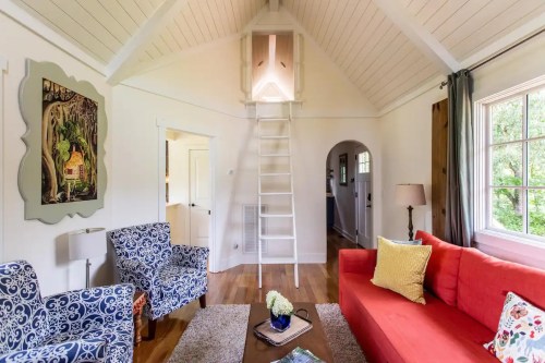 Cozy living room with a red sofa, blue patterned chairs, a ladder to a loft, and large windows for natural light.