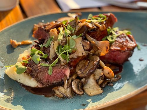A plate of steak topped with sautéed mushrooms and herbs, served on a bed of creamy mashed potatoes.