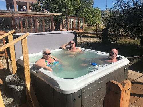 Three people relax in a hot tub on a sunny day, surrounded by trees and a wooden deck.