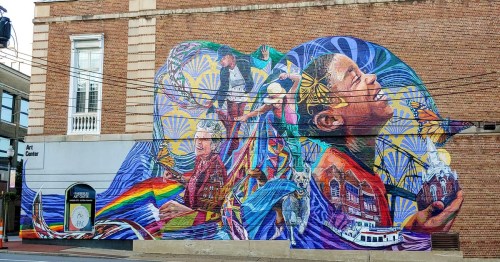 Colorful mural on a building featuring diverse figures, nature, and cultural elements, celebrating community and creativity.