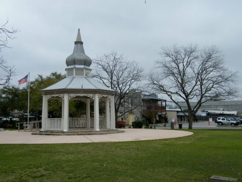 A white gazebo with a pointed roof stands in a park, surrounded by bare trees and a cloudy sky. An American flag is visible.