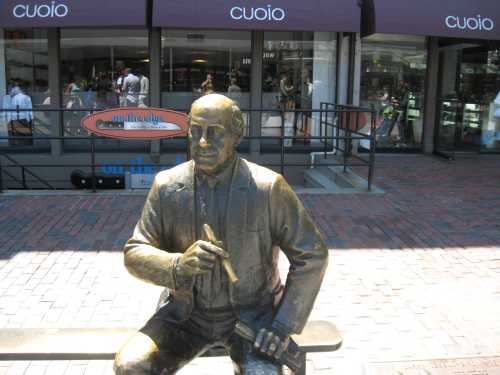 A bronze statue of a seated man holding a pen, with shops in the background.