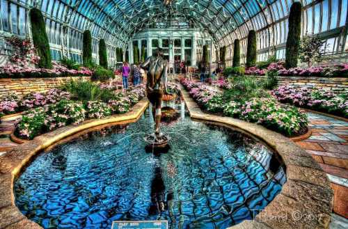 A vibrant greenhouse filled with flowers, featuring a central pond and a statue, surrounded by visitors.