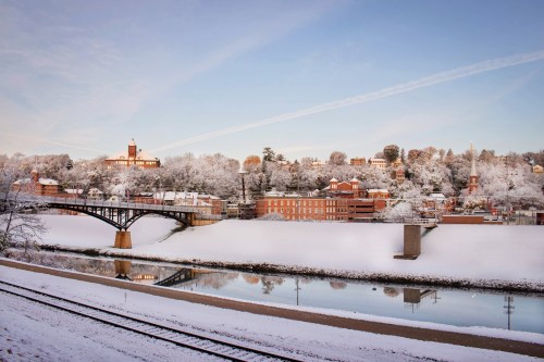 A snowy landscape featuring a river, a bridge, and a charming town with historic buildings under a clear blue sky.