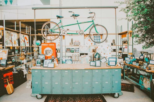 A vintage-style shop display featuring a green tandem bicycle, retro cameras, and various decorative items on shelves.