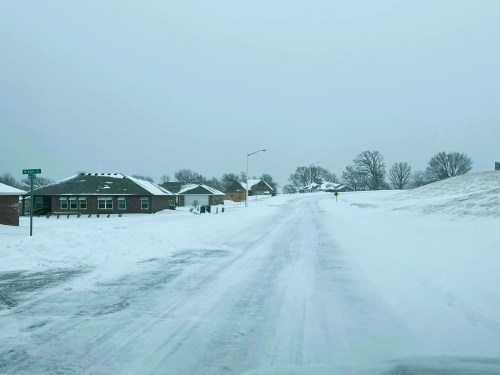 Snow-covered road with houses on either side, under a gray sky, indicating winter weather conditions.