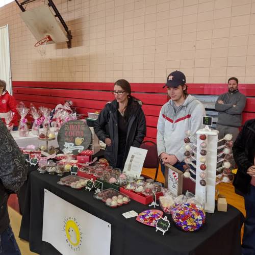 Two vendors at a table displaying various sweets and treats, with customers browsing in a gymnasium setting.