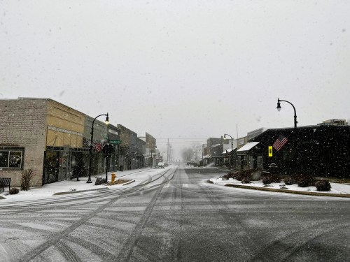 A snowy street scene in a small town, with empty shops and falling snowflakes creating a quiet atmosphere.