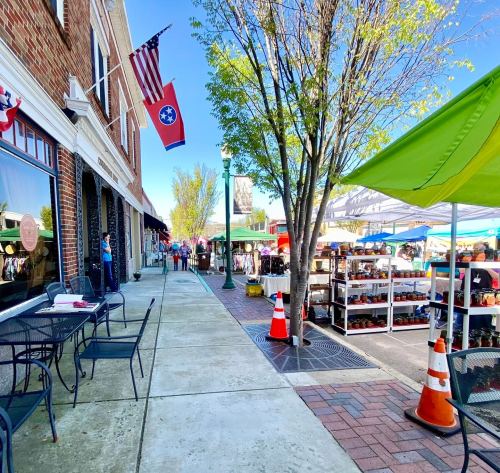 A bustling outdoor market scene with tents, shoppers, and flags in a sunny town setting.