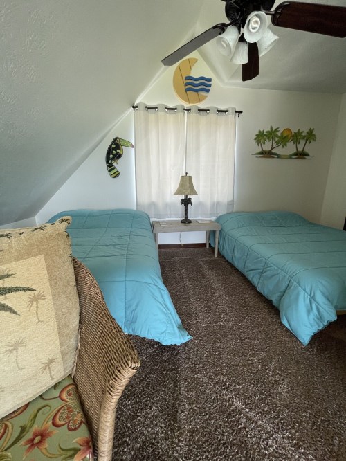 Cozy bedroom with two twin beds, light blue bedding, a lamp, and tropical decor under a sloped ceiling.