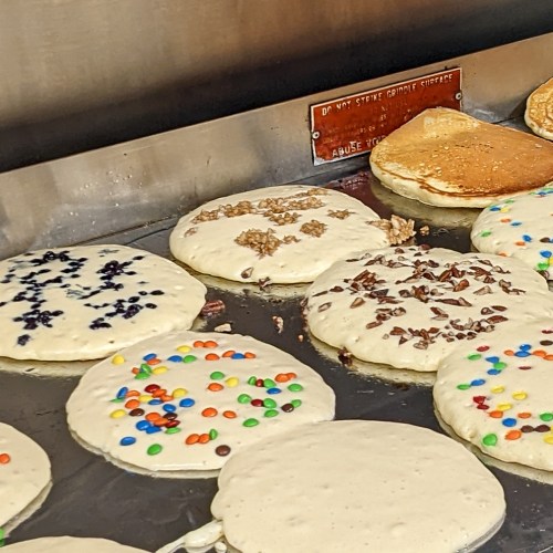 A griddle with various pancakes, some topped with chocolate chips, nuts, and colorful candy pieces.