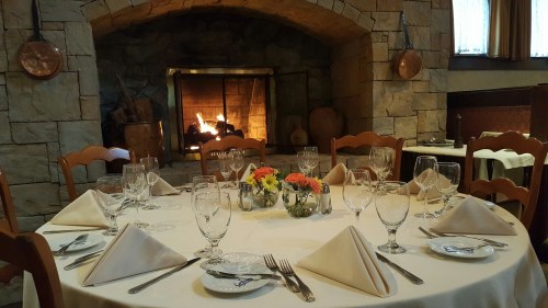 A beautifully set dining table with elegant tableware, surrounded by a cozy stone fireplace and warm decor.