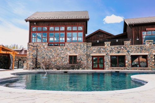 A stone building with large windows overlooks a swimming pool, surrounded by a patio and blue sky.