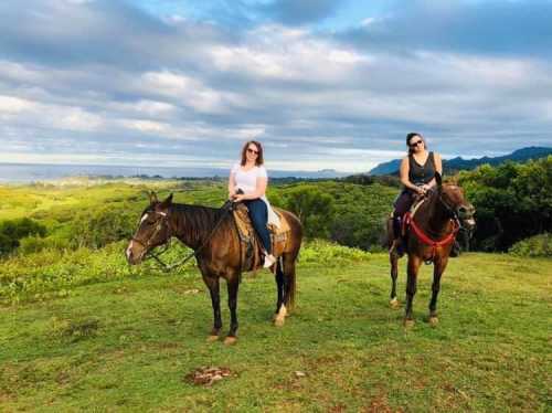 Two women on horseback, enjoying a scenic view of lush greenery and distant hills under a cloudy sky.