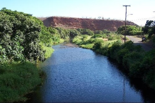 A serene river flows through lush greenery, with a rocky hill in the background and a road alongside.