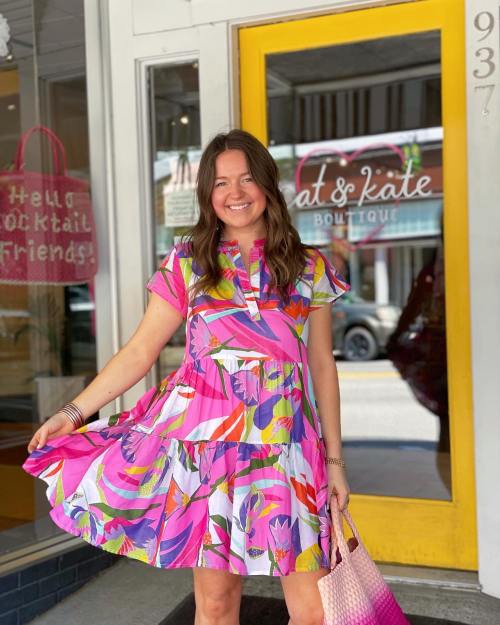 A woman in a colorful floral dress stands outside a boutique, smiling and holding her dress. Bright storefront in the background.