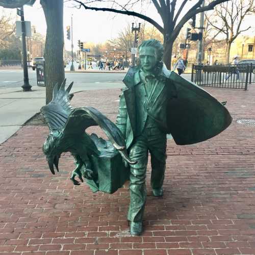 A bronze statue of a man in a coat, holding a bird, set on a brick sidewalk near a street.