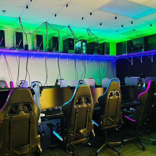 A gaming room with multiple chairs and computers, featuring colorful LED lights and tangled cables on the walls.