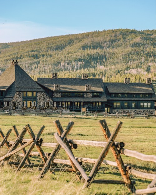 A rustic lodge with a stone facade, surrounded by a wooden fence and lush green fields, set against a mountainous backdrop.
