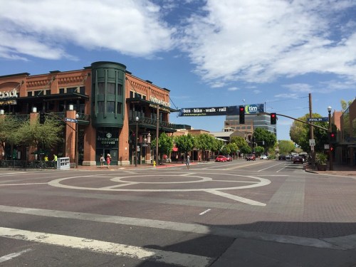 A city intersection with a roundabout, featuring brick buildings, trees, and a blue sky with scattered clouds.