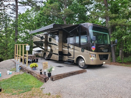 A parked RV with an awning, surrounded by trees and a gravel area, featuring a small wooden deck and potted plants.