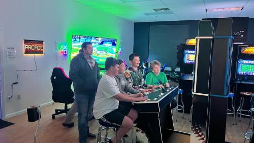 Five kids play arcade games together in a brightly lit gaming room with colorful screens and gaming chairs.