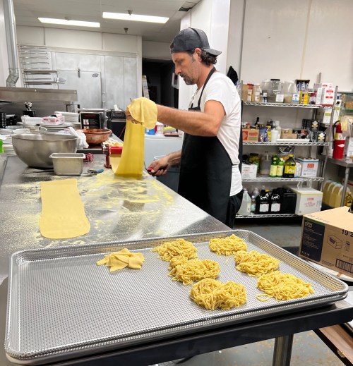 A chef prepares fresh pasta in a kitchen, rolling out dough and shaping noodles on a countertop.