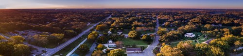 Aerial view of a lush, green landscape with homes and roads under a colorful sunset sky.