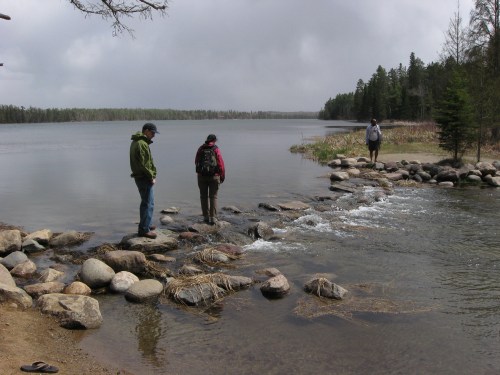 Three hikers navigate a rocky shoreline by a calm lake, surrounded by trees under a cloudy sky.