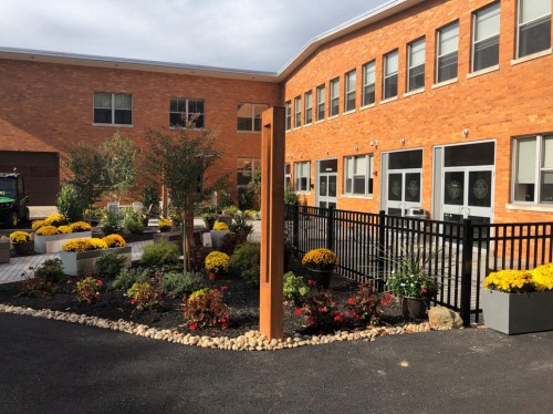 A landscaped courtyard with flowers, trees, and a wooden structure, surrounded by a black fence and brick buildings.