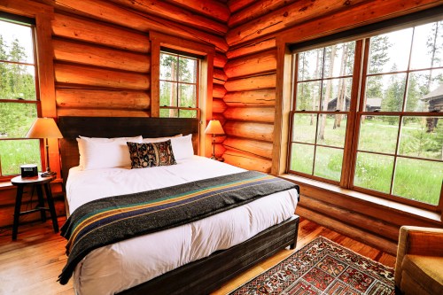 Cozy log cabin bedroom with a large bed, warm lighting, and windows overlooking a green landscape.