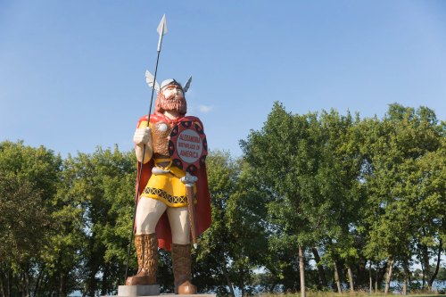 A large, colorful statue of a warrior holding a spear, surrounded by trees and a clear blue sky.