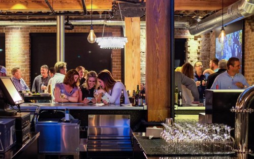 A lively bar scene with people socializing, enjoying drinks, and a stylish interior featuring brick walls and wooden beams.