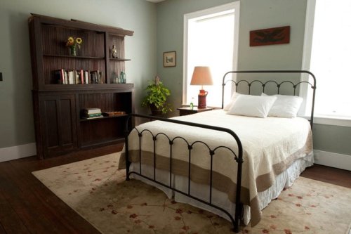 Cozy bedroom featuring a metal bed, bookshelf, lamp, and a plant, with soft lighting and a warm color palette.