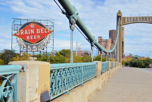 A bridge with turquoise railings leads to a vintage Grain Belt Beer sign against a city skyline and blue sky.