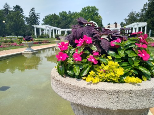 A stone planter filled with vibrant pink and yellow flowers, beside a serene pond and lush greenery in the background.