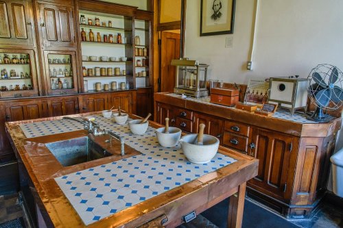 A vintage kitchen with wooden cabinets, a large table with mortar and pestles, and shelves filled with jars and bottles.