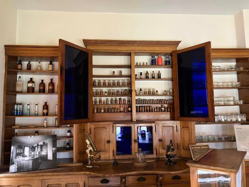 A vintage apothecary cabinet with glass jars and bottles, featuring wooden shelves and a microscope on the counter.