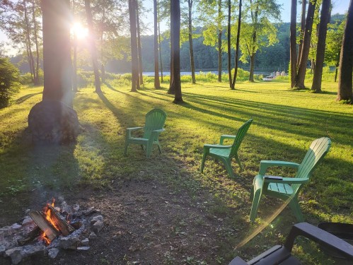 A serene outdoor scene with green chairs around a fire pit, sunlight filtering through tall trees by a lake.
