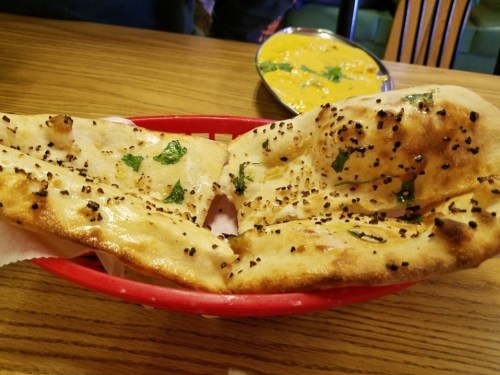 A basket of freshly baked naan bread topped with herbs and spices, served alongside a bowl of yellow curry.