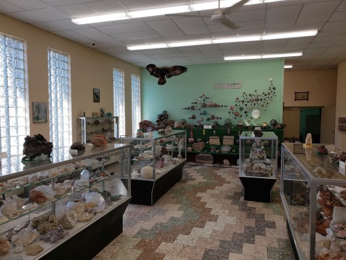 A bright room filled with display cases of various crystals and minerals, with a mural and natural light from windows.