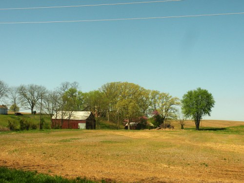 A rural landscape featuring a red barn, trees, and a clear blue sky, with fields in the foreground.