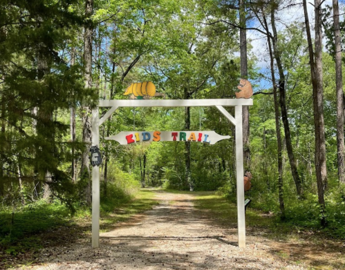 A colorful sign reading "Kids Trail" welcomes visitors to a wooded path surrounded by trees and greenery.