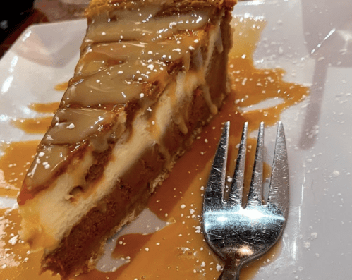 A slice of caramel cheesecake drizzled with sauce, served on a white plate with a fork beside it.