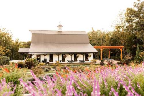 A charming barn-style building surrounded by colorful flowers and greenery, with a wooden pergola nearby.