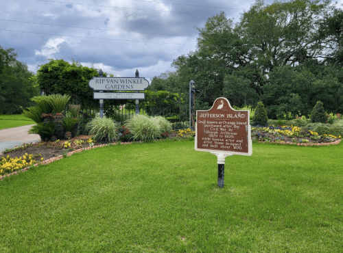Sign for Rip Van Winkle Gardens and Jefferson Island, surrounded by colorful flowers and greenery under a cloudy sky.