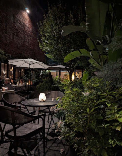 A cozy outdoor dining area with tables, umbrellas, and lush greenery, illuminated by soft candlelight at night.
