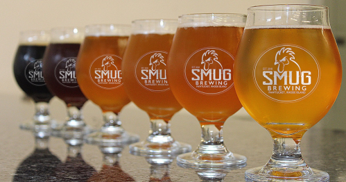 A row of six glasses of beer, each with the SMUG Brewing logo, showcasing different beer colors and styles.